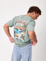 Remera verde claro con gran estampado gráfico en la espalda con motivo de playa, palmeras y la inscripción "Paradise Rush" y "Rusty Sunset Club".