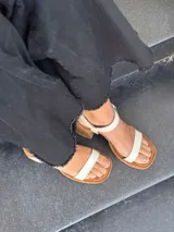 Sandalias de cuero color crudo, con dos tiras anchas sobre el empeine, pulsera al tobillo con hebilla y taco cuadrado de madera.