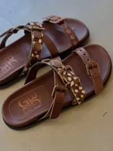 Sandalias de cuero marrón, con diseño de triple tira con hebillas, detalles de tachas y apliques de pelo con estampado animal print.