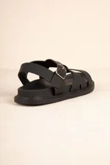 Sandalia baja de tiras cruzadas color camel, con hebilla metálica ajustable en el tobillo y suela negra gruesa.