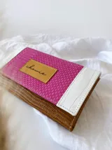 Billetera de cuero con diseño tricolor que combina texturas: cuerpo principal en marrón con textura tipo lagarto, solapa central en fucsia con textura granulada y detalles en blanco con textura tipo cocodrilo. Incluye una etiqueta central de cuero en color tostado con la marca grabada y cierre con broche.