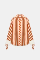 Camisa de manga larga con estampado de rayas verticales en color naranja y crema. Presenta cuello solapa, cierre frontal con botones dorados y puños anchos en color crema con detalle de lazos ajustables.