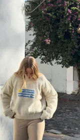Buzo de punto color crema, tejido a mano en lana merino, con diseño de la bandera de Uruguay bordada en el pecho. Presenta cuello redondo y mangas largas con puños acanalados.