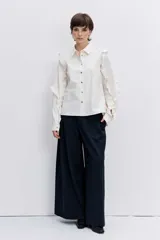 Camisa de color blanco con cuello clásico y cierre frontal de botones. Presenta un diseño de volados decorativos a lo largo de las mangas y los hombros, con puños abotonados.