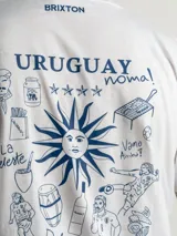 Remera blanca de corte regular con estampado gráfico en la espalda que incluye elementos icónicos de la cultura uruguaya como el sol, un tambor, una parrilla y frases alusivas. Cuenta con cuello redondo y mangas cortas.