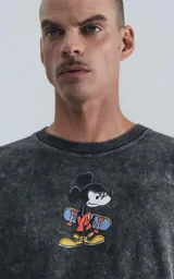 Remera de algodón premium color negro con efecto nevado (acid wash), corte oversize y cuello redondo. Presenta un estampado frontal de Mickey Mouse con una patineta.