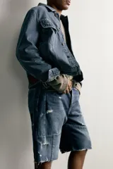 Bermuda baggy fit confeccionada en denim de algodón con lavado azul y efecto rotos en toda la prenda. Presenta diseño de cinco bolsillos, detalles de costuras en las perneras, bajo acabado irregular y cierre frontal con cremallera y botón.