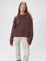 Sweater de tejido de punto trenzado en color marrón, con cuello redondo, puños y ruedo acanalados. Presenta detalles de franjas en color naranja en el cuello y el ruedo, además de apliques de rombos en tonos naranja y verde en las mangas.