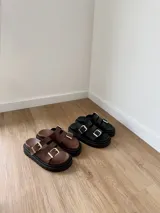 Sandalias marrones de cuero con doble tira y hebillas doradas, sobre base chunky negra.