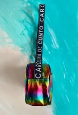 Bandolera Kaira de la línea Ottenta de Carolina De Cunto, confeccionada en material metalizado con acabado multicolor y efecto tornasolado. Cuenta con un bolsillo frontal con broche imantado, cierre superior y una correa larga y ajustable de tela negra con el nombre de la marca en letras blancas.
