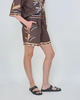 Short color marrón de lino ligero, con cintura elástica con cordón de ajuste, bolsillos laterales y detalles geométricos coloridos en la cintura, dobladillo y estampa de hojas de palmera tropicales.