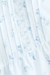 Blusa celeste claro con delicado estampado floretado azul, escote en V pronunciado, mangas cortas tipo mariposa y cierre frontal con botones. Presenta inserciones de encaje y alforzas decorativas en la parte delantera, además de un dobladillo con volados y detalles de encaje.