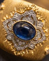 Anillo de bronce con baño de oro, diseño texturizado en relieve con detalles ornamentales y piedra central azul zafiro.