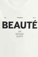 Remera de algodón blanca con cuello redondo, mangas cortas y estampa frontal con la frase "À la recherche de la beauté. Un regard suffit" en tipografía negra.