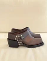 Zueco de cuero marrón con textura envejecida, diseño de punta cuadrada y taco bajo. Presenta una tira trasera con hebilla metálica y detalles de tachas plateadas.