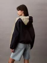 Campera corta con capucha, confeccionada en tejido ripstop de nailon en dos tonos: beige y negro. Tiene corte relaxed, cierre de cremallera y botón de presión, bolsillos con cremallera en la cintura, bajo con cordón elástico, forro de malla transpirable y logo CK bordado en el pecho.