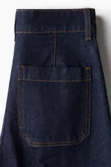 Pantalón de jean azul oscuro, de tiro alto, corte ancho y largo tobillero. Presenta bolsillos delanteros con botón y bolsillos traseros abiertos.