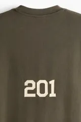 Remera de algodón de punto pesado con efecto lavado, color verde militar. Presenta un corte boxy (holgado) con cuello redondo ribeteado y un pequeño estampado blanco en el pecho con texto y logos.