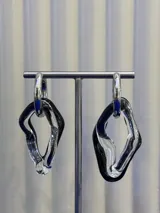 Aros colgantes de diseño irregular, compuestos por una argolla metálica plateada y dos piezas de vidrio soplado entrelazadas en colores negro y transparente.