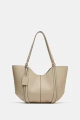 Bolso tipo shopper de hombro en color beige, con diseño estructurado, doble asa y detalle de pespuntes verticales en el cuerpo. Incluye un colgante decorativo tipo etiqueta y cierre imantado.