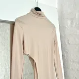 Top de lycra color beige, con cuello alto y mangas largas. Presenta un corte asimétrico en la parte inferior.