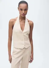 Chaleco sastrero de lino color beige, con cuello halter pronunciado y cierre frontal de cuatro botones. Presenta bolsillos ribeteados simulados en el frente y dobladillo en punta.