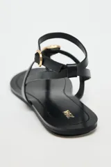 Sandalia plana negra con tira para el dedo y tira tobillera ajustable con hebilla dorada. Presenta un adorno grande de pieza metálica dorada con textura orgánica sobre el empeine.