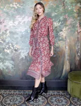 Vestido camisero midi de gasa estampada con flores rojas y blancas sobre fondo marrón. Tiene cuello con solapa, abertura central con botones, mangas largas y lazo de ajuste en la cintura.