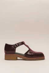 Zapato tipo guillermina de cuero charol color marrón oscuro, con tira T y hebilla ajustable en el lateral. Posee puntera cuadrada y suela de goma color caramelo con taco bajo.