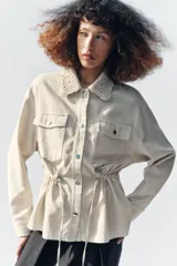 Chaqueta estilo safari color beige, con cuello de solapa tejido en crochet, manga larga, bolsillos delanteros con solapa y cintura ajustable con cordones. Cierre frontal con botones metálicos a presión.