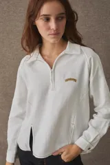 T-shirt de manga larga confeccionada en mezcla de algodón y lino, color blanco, con cuello camisero, cierre frontal parcial con cremallera, puños abotonados y aberturas laterales en el bajo. Cuenta con un pequeño bordado distintivo en el pecho.