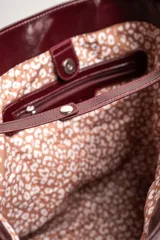 Bolso tipo tote de gran tamaño en color bordó con acabado brillante. Cuenta con doble asa de hombro y placa metálica decorativa con logo en la parte frontal.