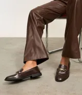 Zapato mocasín femenino color marrón oscuro, confeccionado en poliuretano, con detalle metálico dorado en forma de lazo en el empeine y tacón bajo.