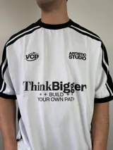 Remera de diseño estilo jersey deportivo con calce oversize, color blanco con mangas cortas negras y detalles de franjas blancas en los hombros. Cuenta con estampado frontal con texto y logotipos.