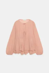 Blusa de viscosa color rosado con cuello redondo, manga larga abullonada con puños elásticos y cierre frontal con lazadas. Presenta un diseño con volantes en el bajo y corte asimétrico.