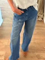 Pantalón de jean azul con corte holgado y detalle de tres cruces bordadas en el muslo derecho. Presenta costuras reforzadas y cierre frontal con botón metálico.