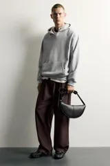 Bolso de hombro pequeño color negro con acabado liso, diseño semilunar y costura central. Cuenta con compartimento principal con cierre de cremallera, bolsillo interior con cierre y asa de hombro ajustable con hebilla.