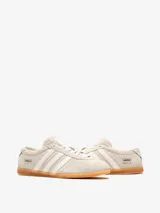 Championes Adidas Gazelle Lo Pro W de perfil bajo, confeccionados en gamuza color gris claro, con las tres tiras características en tono crudo y suela de goma translúcida. Incluye el nombre del modelo y la marca en letras doradas en el lateral.