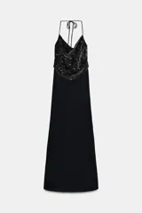 Vestido largo negro de corte recto, con escote halter ajustable mediante lazada y escote fluído. Presenta un detalle de tejido superpuesto en terciopelo negro con aplicación de abalorios y lentejuelas en la parte superior.