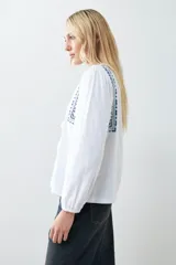 Remera blanca de manga larga con detalles bordados en tonos azules y verdes en el pecho y hombros. Presenta un escote en V con cordones ajustables terminados en borlas y puños elásticos.