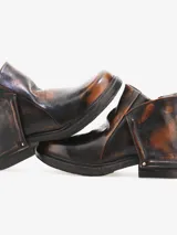 Bota corta de cuero bovino negro con efecto desgastado y envejecido, con detalles en gris claro. Presenta un diseño con solapa frontal que se ajusta con un broche y una cremallera lateral expuesta en el talón. Posee un taco bajo y cuadrado.