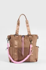 Cartera tipo shopper color marrón, con compartimento principal con cierre, bolsillos laterales, bolsillo frontal de red y cordones decorativos en color rosado. Cuenta con correas dobles de hombro y una correa extraíble de cuerda trenzada en color rosado y blanco.
