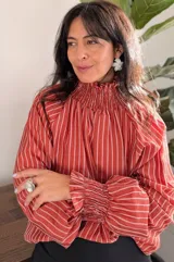Blusa de tela liviana con diseño de rayas verticales en color terracota y blanco. Presenta cuello alto con detalle de smock, mangas largas con volumen y puños también con detalle de smock.