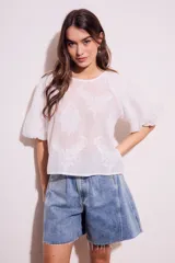 Blusa blanca traslúcida con estampado floral en relieve, cuello redondo y mangas globo cortas.