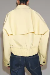 Chaqueta corta confeccionada en mezcla de algodón color amarillo claro, con cuello de solapa, manga larga, bolsillos delanteros y cierre frontal con botones visibles en los extremos.