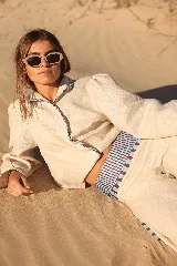 Camisa de lino color beige con mangas globo y detalles de vivos estampados a rayas en azul y marrón. Presenta cierre frontal con botones y diseño anudable en la cintura.