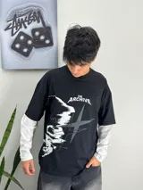 Remera negra de manga larga con diseño en capas que simula una remera de manga corta negra superpuesta sobre una manga larga blanca. Presenta estampa frontal en blanco y gris con la inscripción "The Archive" y gráficos abstractos.