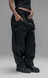 Pantalón de estilo utilitario y corte oversize, confeccionado en textil técnico liviano. Presenta cintura elástica con cordón ajustable, bolsillos laterales envolventes de gran tamaño con cordones y tancas de ajuste, y elásticos regulables en los tobillos.