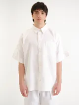 Camisa blanca de manga corta, con cuello clásico, cierre frontal con botones y un bolsillo aplicado en el pecho.