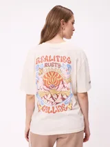 Remera de algodón con corte holgado, color crema, con estampado gráfico en la espalda que incluye un sol, montañas y mariposas con la frase Realities Rusty Collide.